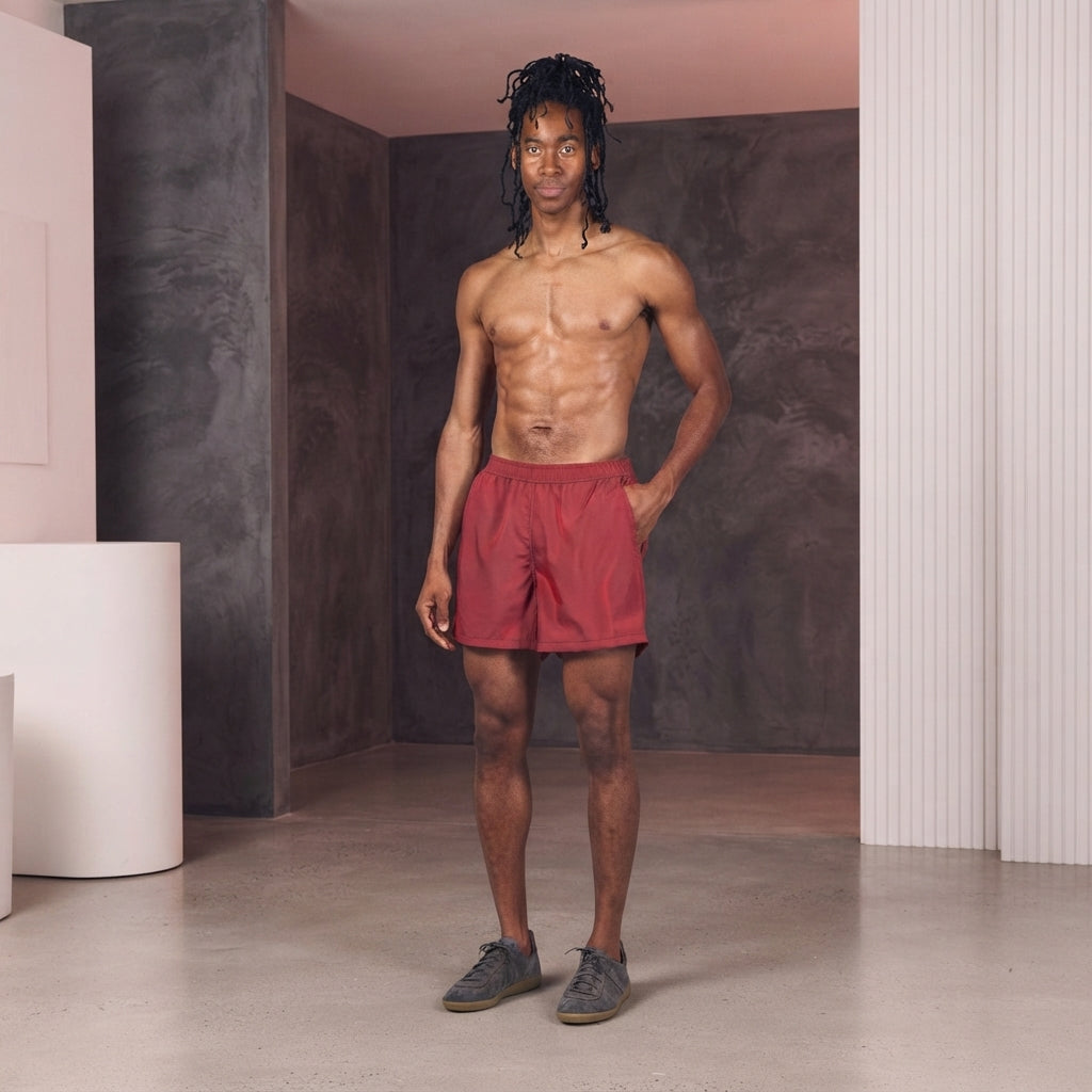 A model wearing a crimson iridescent trunk in a minimalist studio with charcoal and white accents.